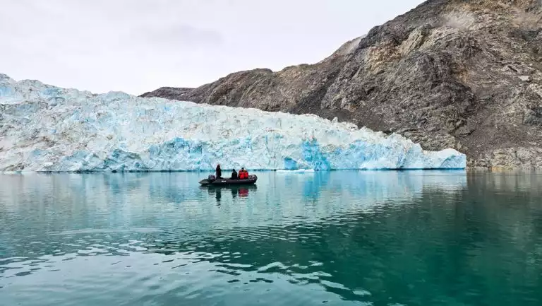 Zodiac boat with East Greenland cruise guests idles in glassy teal water beside large, chunky blue glacier & dark cliffs.