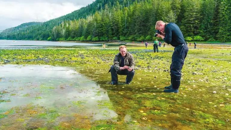 Man photographing the green mossy ocean while his friend kneals next to him in front of lush green forest