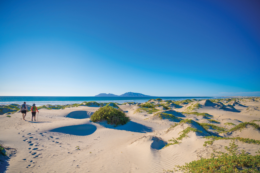 two hikers walk through the sandy landcape and out to the beach head on a bright sunny day