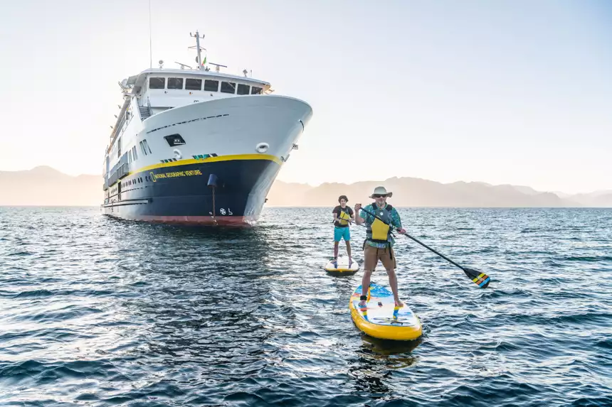 Father and son go out for a paddle board on hazy day next to small cruise ship national geographic venture 