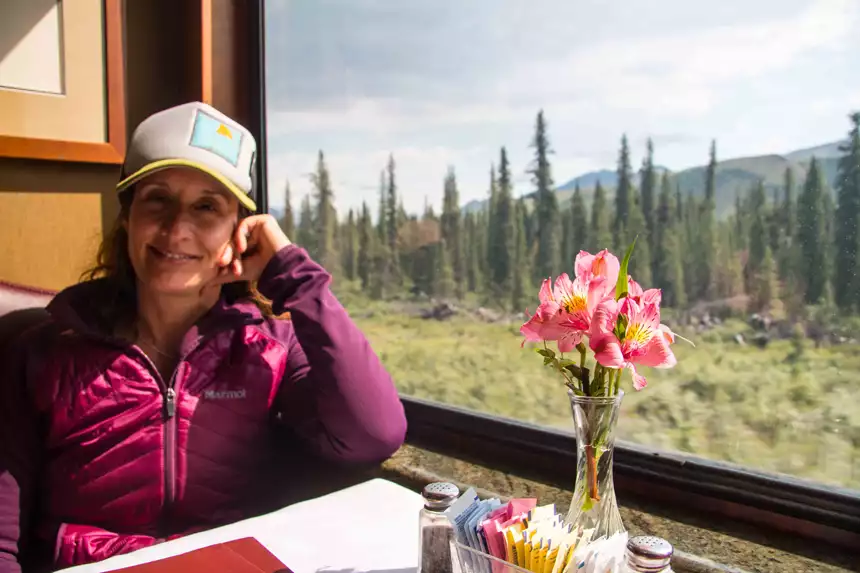 Woman in berry-colored coat sits with head propped on hand at table with pink flowers & view window on Alaska Railroad dining car.