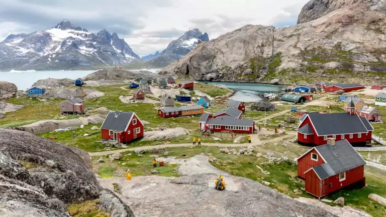 Iceland to Greenland traveler in yellow jacket sits on rock overlooking small arctic town with red buildings by the sea.