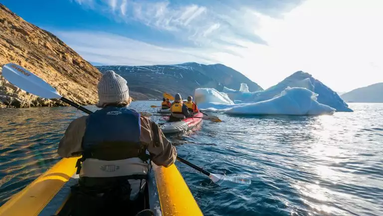 Man dressed warmly paddles front of yellow inflatable kayak beside tan rock cliffs & blue floating icebergs in Greenland.