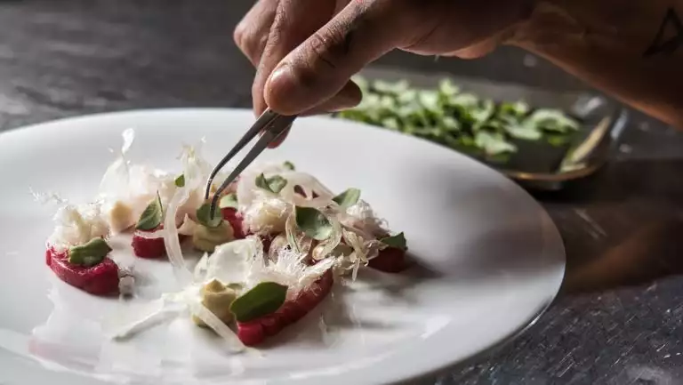 Chef's hand holds tweezers & places pieces of craft vegetables on a white plate during an Iceland to Greenland cruise.