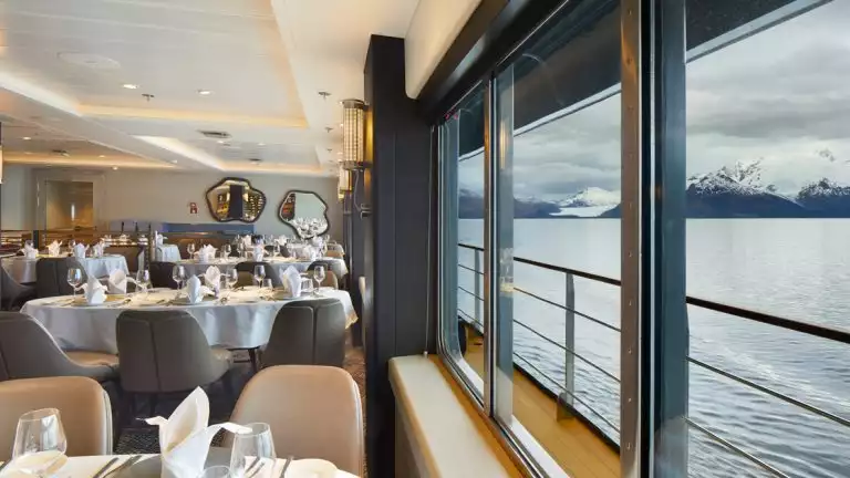 Restaurant on Magellan Explorer ship with grey & tan leather chairs around white table clothed tables & view windows.