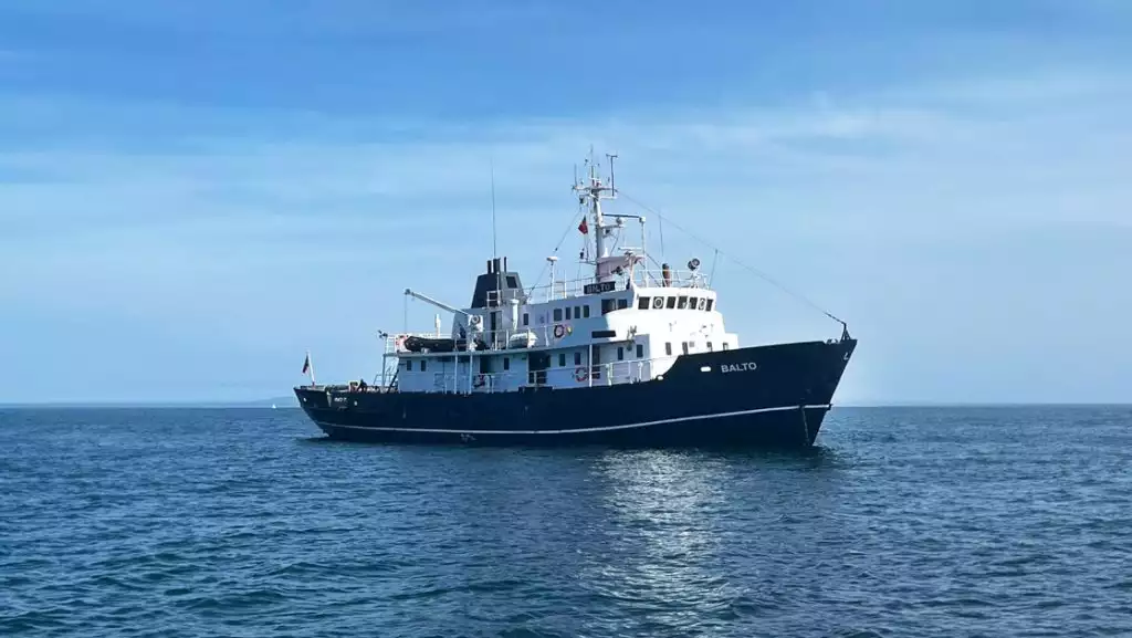 MS Balto yacht with dark blue hull & 2 white upper decks sits in calm Arctic ocean on a sunny day with blue skies.