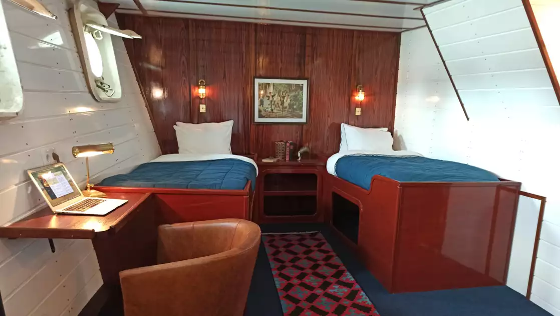 Superior cabin on MS Balto with 2 fixed twin beds, white & wood walls, 2 portholes, runner rug, desk & leather chair.