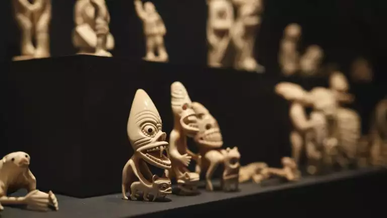 Small beige carved characters with peaked heads, large eyes & wide mouths sit on display in a South Greenland museum.