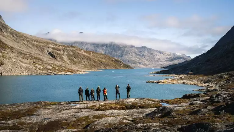 Small group of hikers stand atop a rocky outcrop overlooking a fjord with deep blue water flanked by rocky mountains.