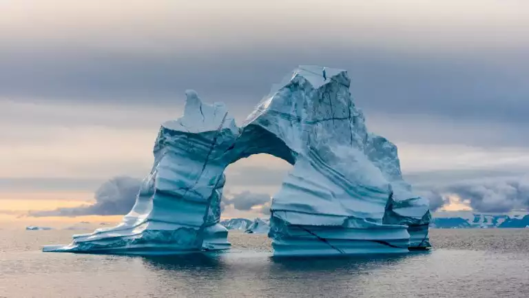 Large iceberg with natural arch & striations of blue & black rises out of calm sea, seen on a South Greenland cruise.