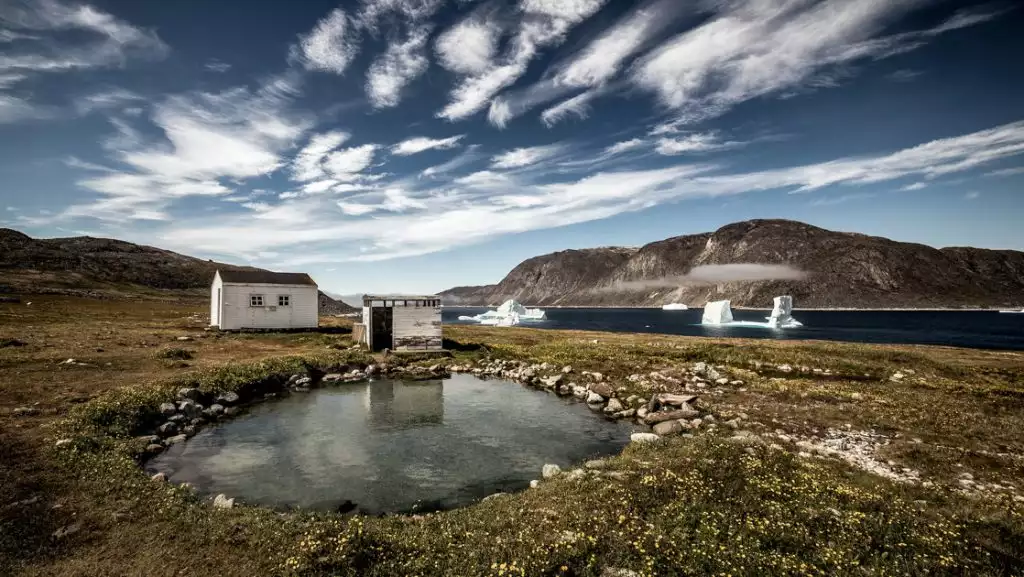 Natural hot spring with weathered small building beside fjord with large white icebergs & mountains behind in Greenland.