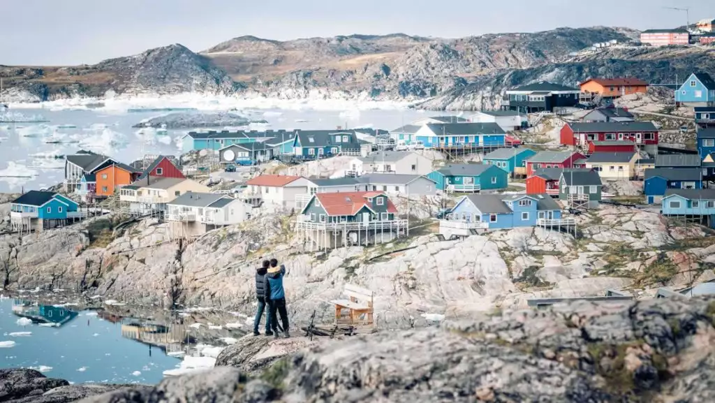 2 West Greenland travelers stand on a rocky hillside overlooking a small town with colorful buildings beside an icy bay.