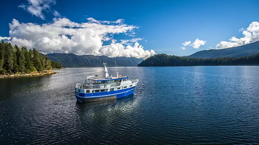 A small blue hulled Alaska yacht charter ship seen at anchor in a still cove with blue skies