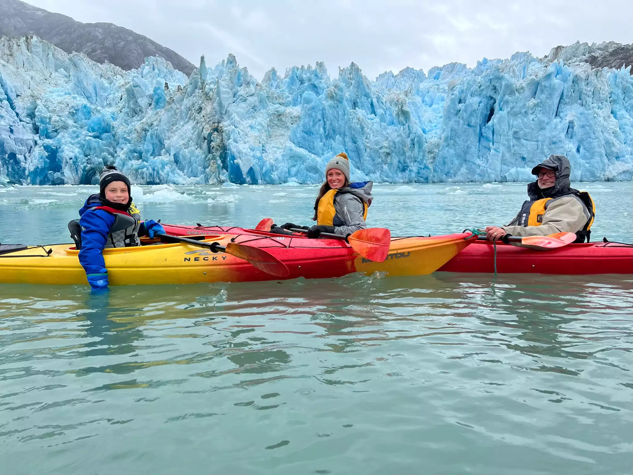 A family of two adults and one child in a blue jacket sit in kayaks in front of a big blue glacier