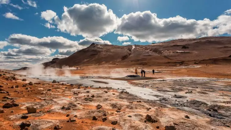 Iceland travelers walk over Hverarondor Hverir geothermal area with its multicolored, cracked mud & steam under clouds.