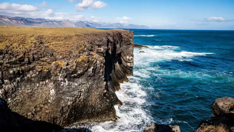 Scenic Iceland coast with golden tundra, tan cliffs & turquoise sea with large swells hitting the shore on a sunny day.