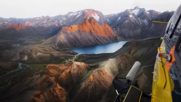 View from yellow helicopter as it flies over jagged peaks with green valleys & a small lake in Iceland.