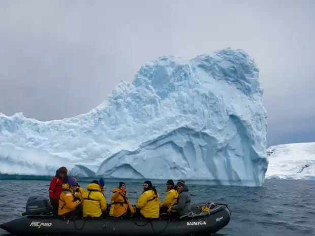 A zodiac full of travelers in yellow jackets passes by a big iceberg in Antarctica