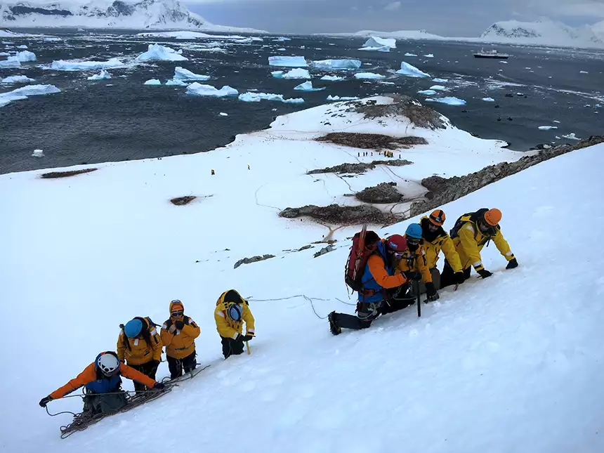 People on a mountaineering excursion in Antarctica climbing uphill on snow with ice picks and ropes with their small ship in the background. 