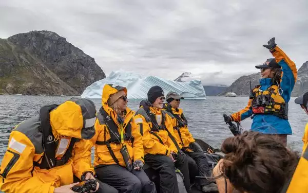 A Quark expedition guide raises a hand while driving a zodiac and speaking to a group of travelers in yellow jackets with an iceberg behind them