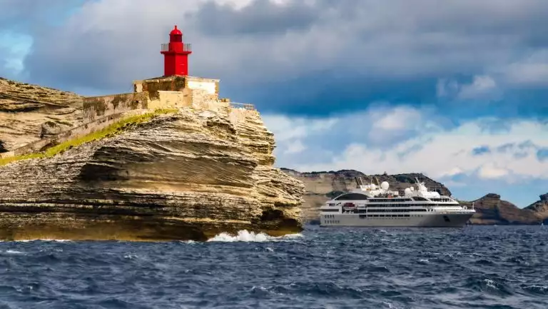 Modern gray & white expedition ship sits by tan cliffs under a red lighthouse during a Mediterranean solar eclipse cruise.