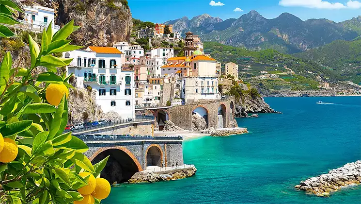 Lemon trees look onto turquoise water, sea walls & historic seaside town on a sunny day in Italy.