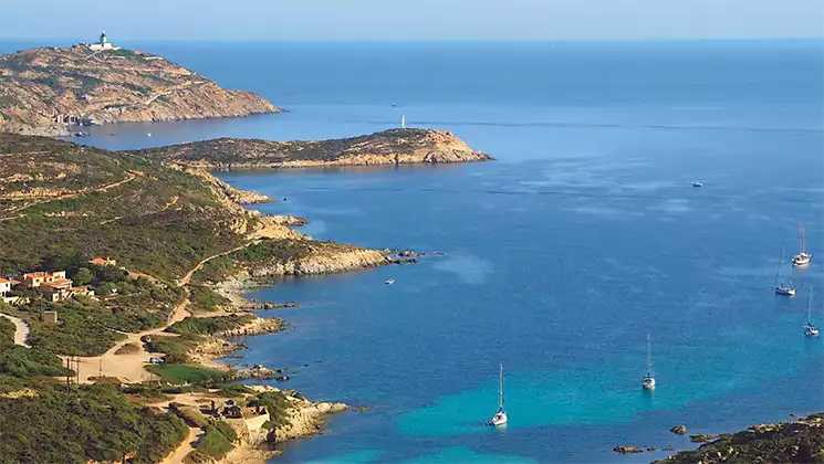 Aerial view of La Revellata, Italy with tan shoreline, white-sand beach & green hills by open ocean with sailboats at anchor.