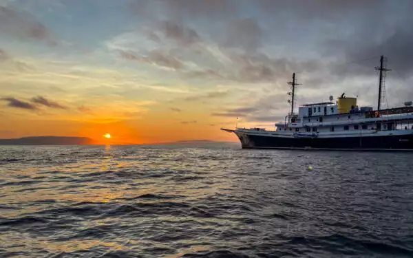 Small ship cruise pointing into the sun as it shines bright orange and yellow over the calm waters of Galapagos