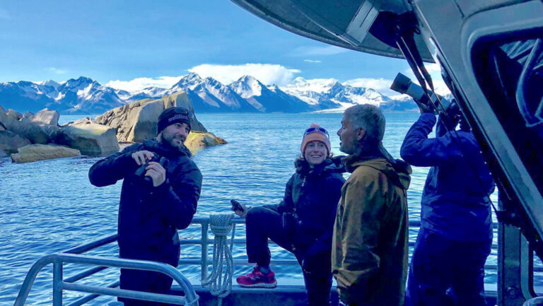 Small group of travelers on an Alaska hiking tour stand on bow of small boat talking & viewing scenic snowy peaks.