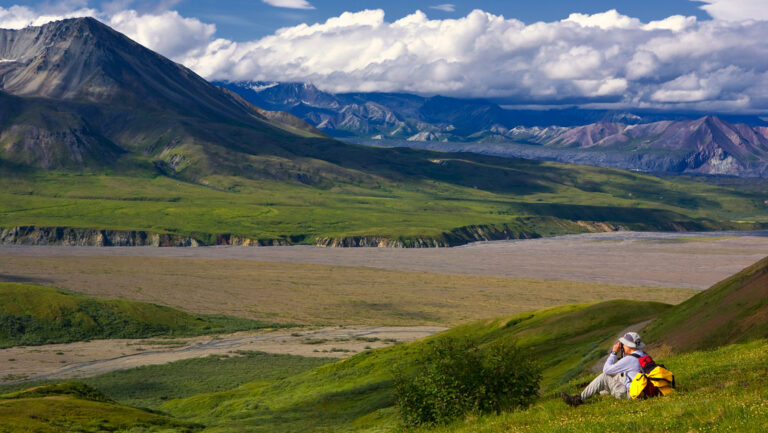 Male hiker with backpack & sun hat sits on a grassy hillside & looks at mountains & valleys on an Alaska hiking vacation.