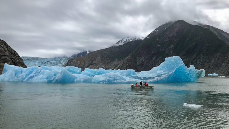 Small tender boat with Alaskan Story charter cruise guests slowly motors past massive blue iceberg chunks on a cloudy day.