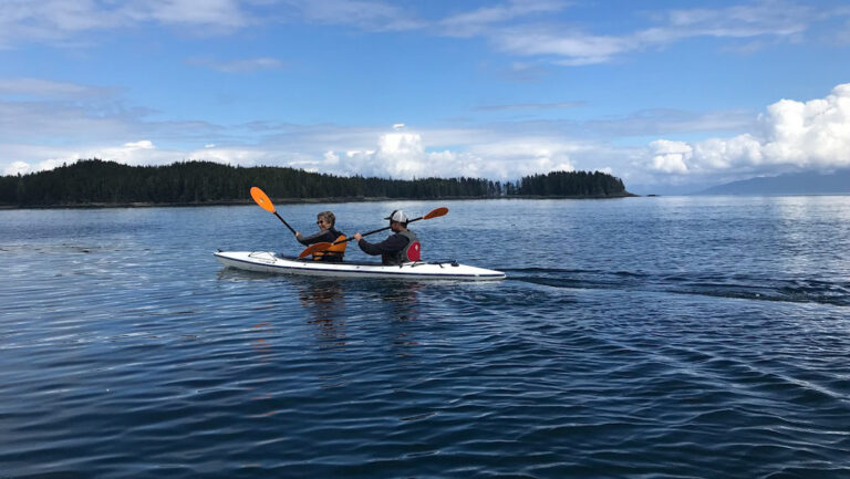Tandem kayakers paddle a white boat in calm sea backed by green hills on a sunny day in Alaska.