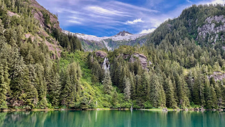 Scenic southeast Alaska with verdant mountains with granite peeking out & a waterfall spilling into turquoise sea.
