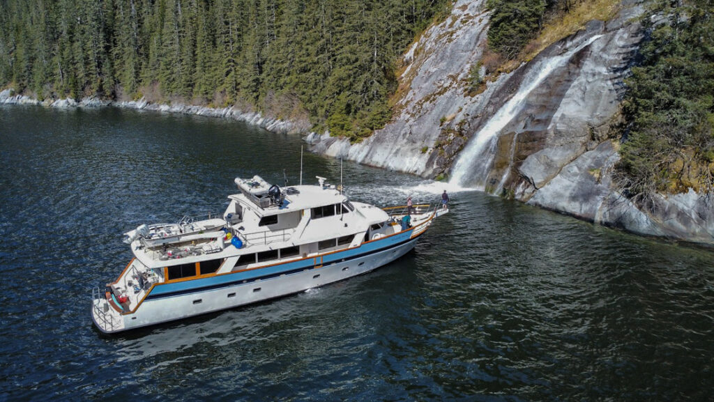 Aerial view of Alaskan Story yacht with blue & white exterior sitting in calm water in front of water cascading over rock.