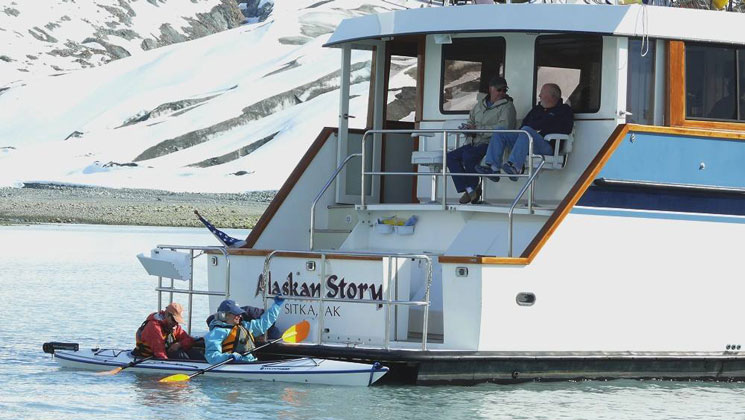 Aft of Alaskan Story yacht with tandem kayakers alongside waterline swim step as 2 guests sit in seats above.