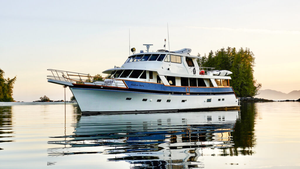 Alaskan Story yacht with 3 decks, kayaks, paddleboards & blue & white exterior paint sits in glassy water at dusk.