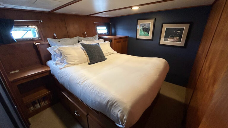 Walkaround queen bed in white linens in cabin with blue & wood walls & 2 rectangular portholes on the Alaskan Story yacht.