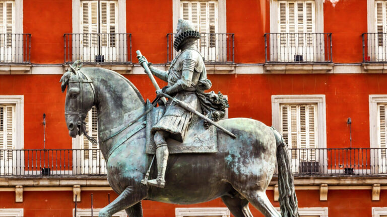 statue of soldier riding a horse with a weapon in hand infront of bright red building with white balconys