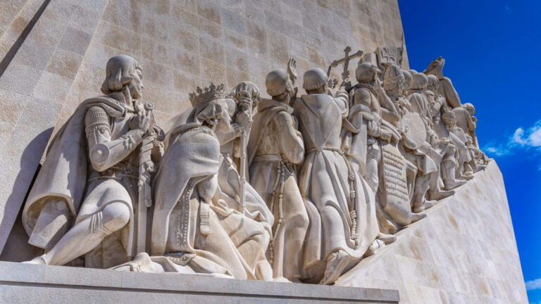 Stone statue on the side of a historic building showing men and women walking up stairs to the bright blue sky