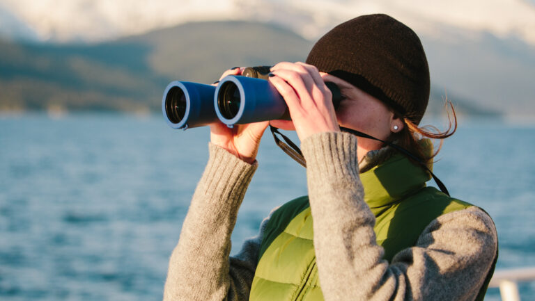 Woman in black knit cap & green puffy vest holds blue binoculars up to look at wildlife on a Gulf of Alaska cruise.