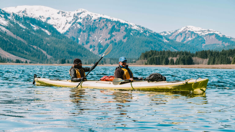 Tandem kayakers paddle a yellow boat in calm water with snowcapped peaks behind on a sunny day of a Gulf of Alaska cruise.