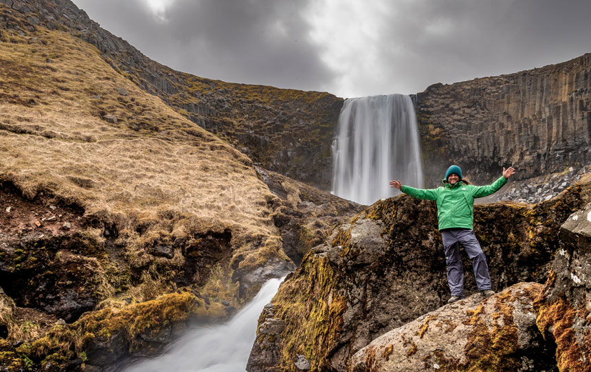 Man in green jacket, knitted blue hat & gray pants stands in front of big waterfall among rocks in Iceland.