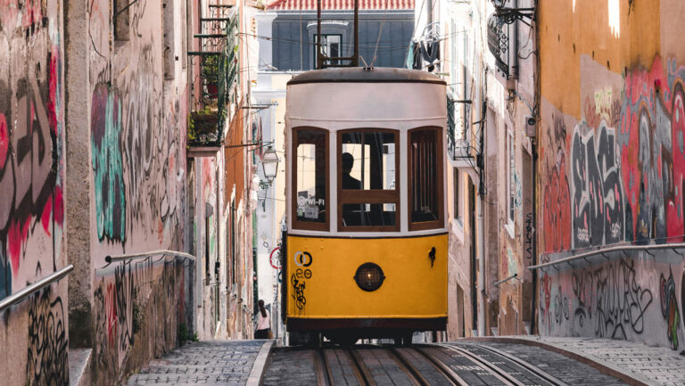 Yellow & white cable car climbs stone street between two graffitied buildings in Lisbon, Portugal, seen on the Med Feast cruise.