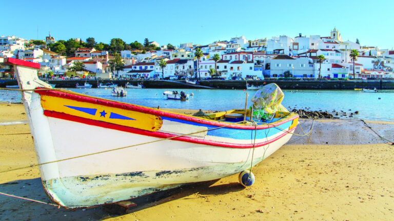 Small wooden boat pulled onto the beachy shore with beautiful white costal town behind
