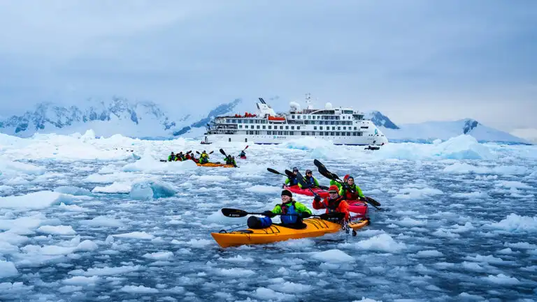 group of kayakers in brightly colored kayaks paddle through dense iceberg field. Greg Mortimer expedition ship anchored behind
