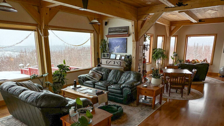 Great Room at Denali Overlook Inn with tall wood ceilings, walls of windows, 2 overstuffed green couches & coffee tables.