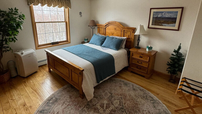 Queen-sized wooden sleigh bed with teal & white linens beside view window & atop wood floors at Denali Overlook Inn in Alaska.