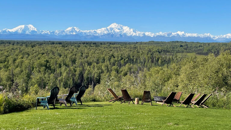 Large green lawn with Adirondack chairs around a firepit overlooking forest and snowcapped Mt. Denali in the distance.
