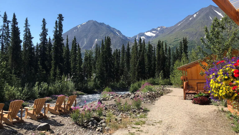 Wooden McKinley Creekside Cabins & Adirondack chairs sit on the riverbank under tall green peaks and beside forest in Alaska.
