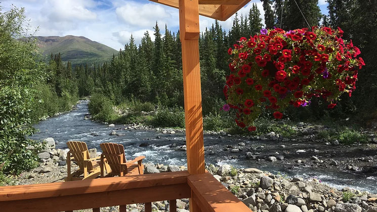 Wooden deck with flowering hanging basket & 2 Adirondack chairs sit creekside among rocks & green forest in the sun in Alaska.